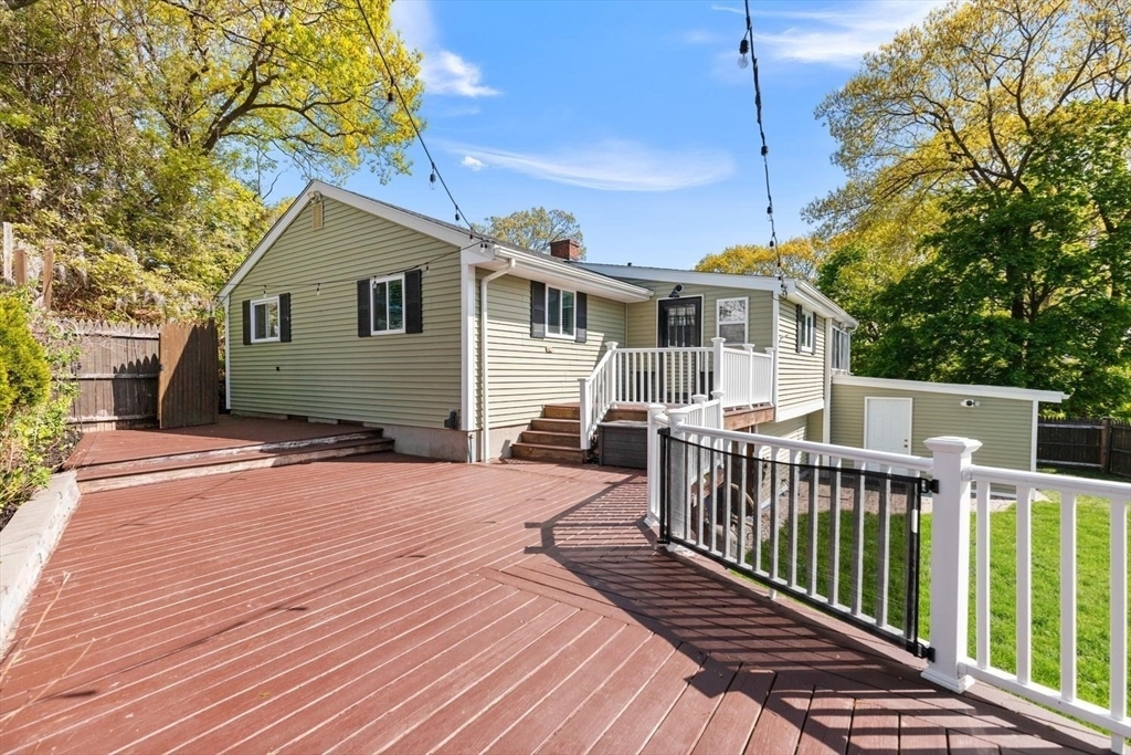 The back of 53 Longhill Rd seen from its multi-level wood deck, sage siding and white railings above a sloped lawn ringed by mature trees under a blue sky