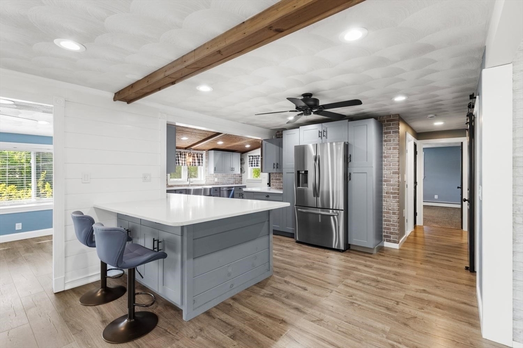 Renovated kitchen at 53 Longhill Rd with a white quartz island, two gray barstools, gray shaker cabinets, a stainless French-door refrigerator, a reclaimed-wood ceiling beam, and a brick column