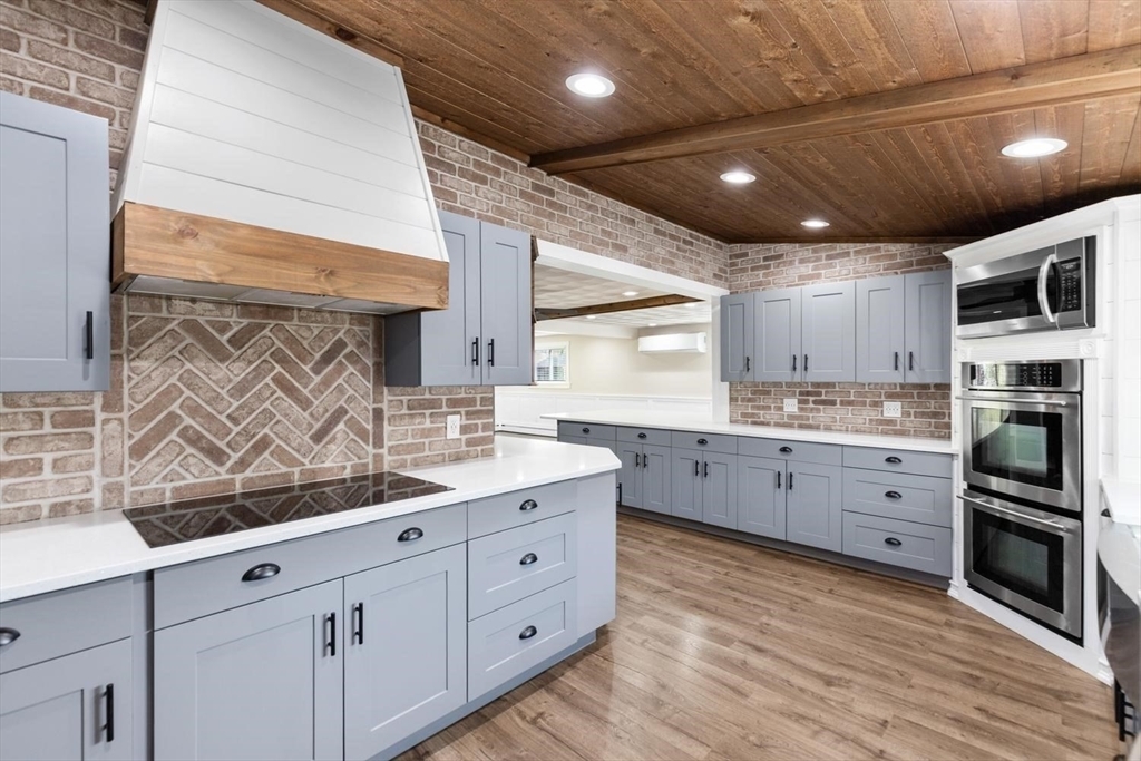 Kitchen detail showing a herringbone brick backsplash behind a black induction cooktop, a white range hood with a wood band, gray cabinets, and a plank wood ceiling