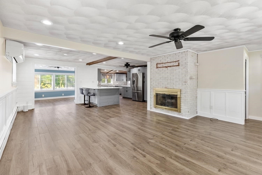 Living room at 53 Longhill Rd with a white-painted brick fireplace and brass surround, a ceiling fan, wood-look floors, and an open sightline through to the kitchen