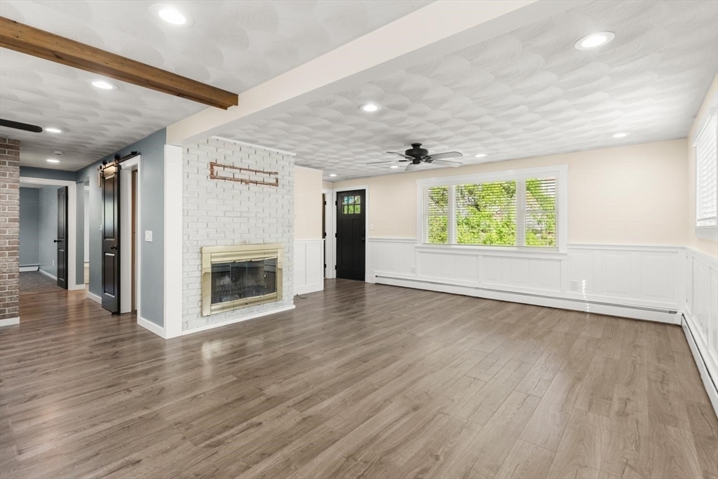 Open living and dining area at 53 Longhill Rd with a white-painted brick fireplace, a black glass-paned front door, a large picture window with plantation shutters, white wainscoting, and wood-look floors