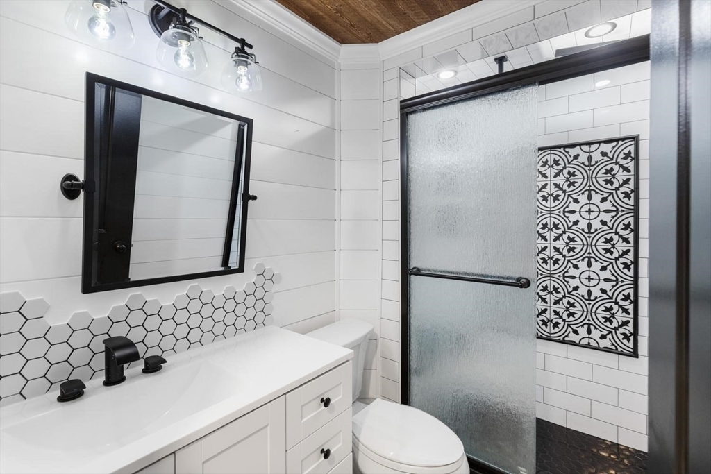 Renovated full bath with white shiplap walls, a black-framed mirror, a three-globe sconce, white hex tile, and a glass shower with a black-and-white patterned accent tile under a wood plank ceiling