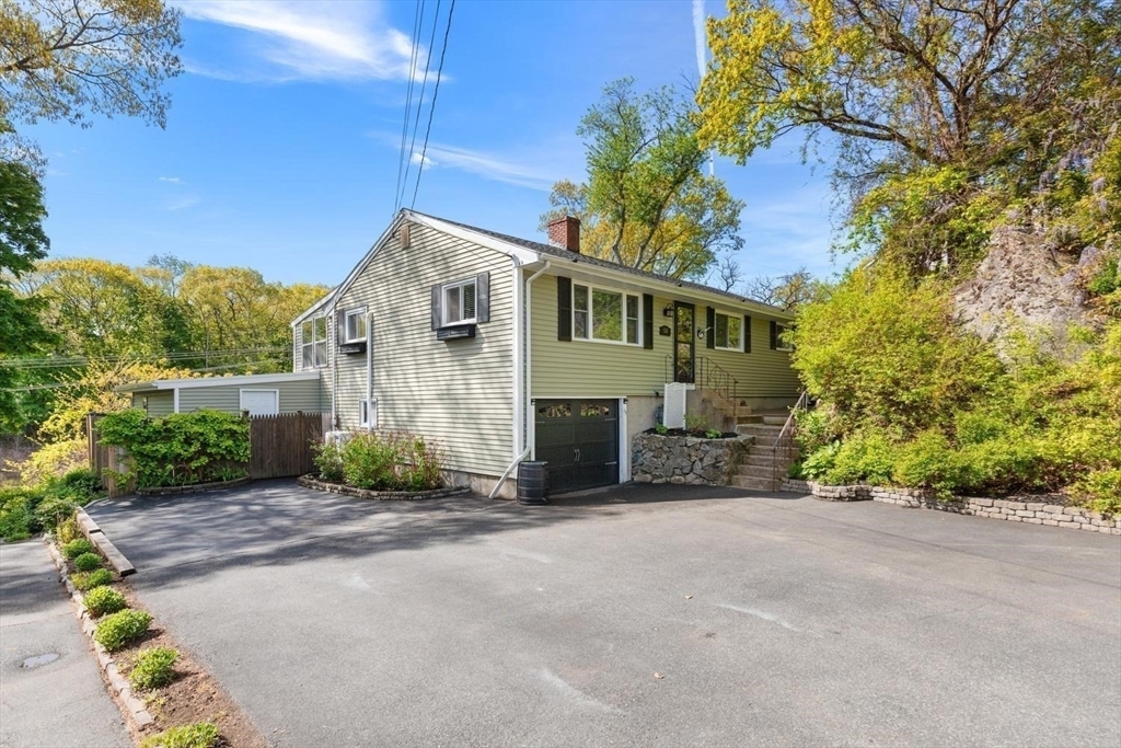 Front three-quarter view of 53 Longhill Rd, a renovated single-level ranch with sage clapboard siding and black shutters, set against a wooded hillside in Lynn, Massachusetts